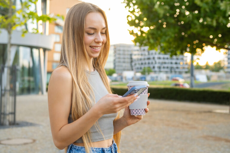 blonde hair woman smiles phone screen texting message standing street sunset scaled